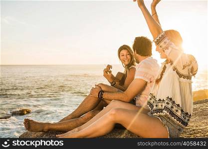 Three mid adults sitting on rocks playing acoustic guitar at sunset, Cape Town, South Africa