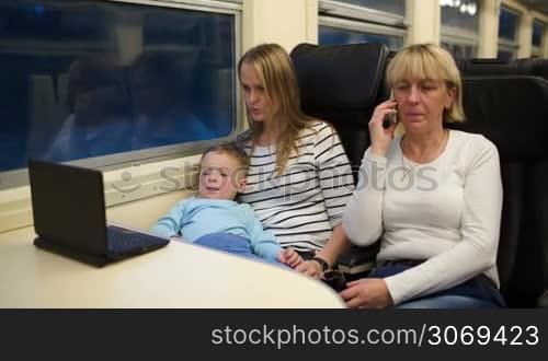 Three passengers in th train in the evening. Mother and son watching movie on computer, grandmother having a phone talk