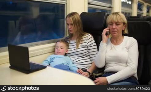 Three passengers in th train in the evening. Mother and son watching movie on computer, grandmother having a phone talk