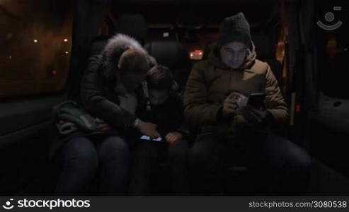Three passengers traveling by minibus in late evening. Mother and son entertaining with smart phone and man passing the time with browsing on mobile