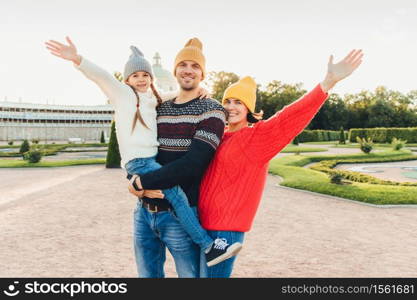 Three people have good time together: mother, father and small daughter stand next to each other, walk outoors, wave with hands, have happy expressions. People, facial expressions and emotions
