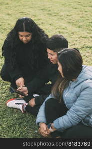 three people of hispanic-latino ethnicity sitting on the green grass of a park, with smart device. relaxed and comfortable as they entertain themselves with their screens, browsing social networks, dressed in casual clothes and their faces suggest satisfaction with their activity. it conveys a sense of modernity and connectivity that is common in today’s digital age.. three people of Hispanic-Latino ethnicity, sitting on the ground in the park with smart devices