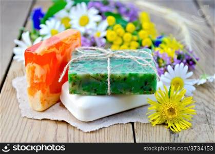 Three pieces of homemade soap on a piece of paper, chamomile flowers, tansy, elecampane on the background of wooden boards