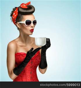 Three-quarter portrait of glamourous pinup girl wearing vintage gloves and red ribbon in her hair, holding a cup of hot coffee or tea and cooling it.