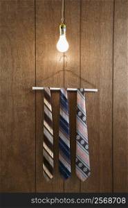 Three retro ties hanging on a wire hanger against wood paneling with a glowing lightbulb hanging down from above.