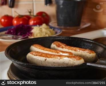 Three sausages fried in cast iron pan, tomatoes and sauerkraut on background