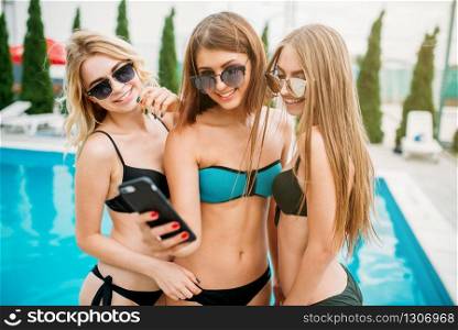 Three sexy girls in swimsuits and sunglasses makes selfie near the swimming pool. Resort holidays. Tanned women on summer holidays. Sexy girls in swimsuits makes selfie near the pool