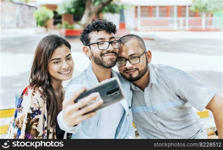 Three smiling friends sitting on a bench taking a selfie. Front view of three happy friends taking a selfie while sitting on a bench. Three friends taking a selfie on a park bench