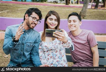 Three smiling friends taking a selfie sitting in the park, Three smiling friends sitting in the park taking a selfie. Front view of three happy friends taking a selfie while sitting on a bench