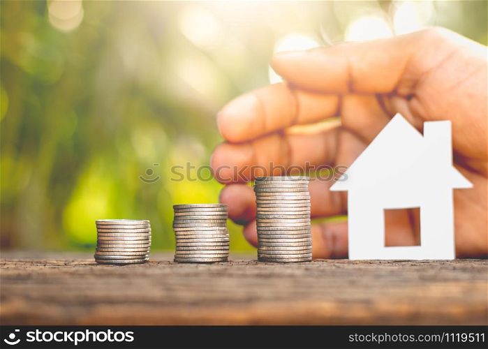 Three stacked coins on old wooden floor. As the men&rsquo;s hands are wrapped around the small white paper house with the morning sunlight shining.