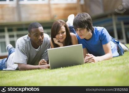 Three students lying outdoors on lawn with laptop
