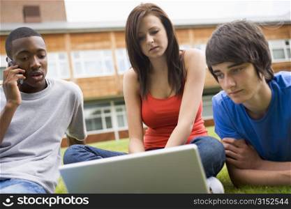 Three students outdoors on lawn with laptop one on cellular phone