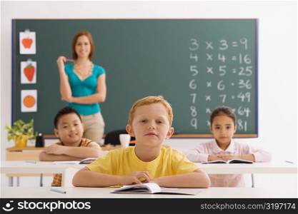 Three students sitting in a classroom with their teacher standing in the background