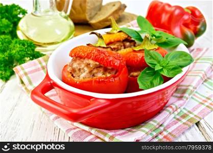 Three sweet peppers stuffed with meat and rice with basil leaves in a red roasting pan on a napkin, parsley, oil, bread on the background light wooden boards