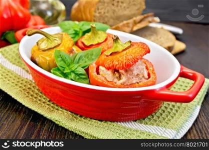 Three sweet peppers stuffed with meat and rice with basil leaves in red roasting pan on a green napkin, bread on a dark wooden board