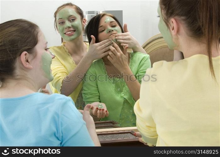 Three teen girls in bedroom mirror putting on facial mask. Focus on reflection of girl in yellow.