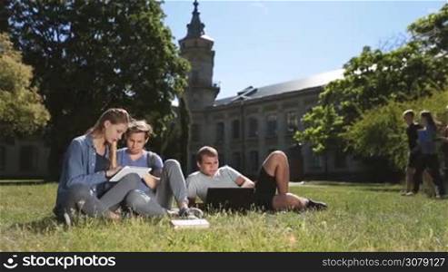 Three teenage hipster friends studying on campus lawn, using laptop and digital tablet pc while sitting on green grass outside university building. College students learning together with the help of modern technology devices. Dolly shot.
