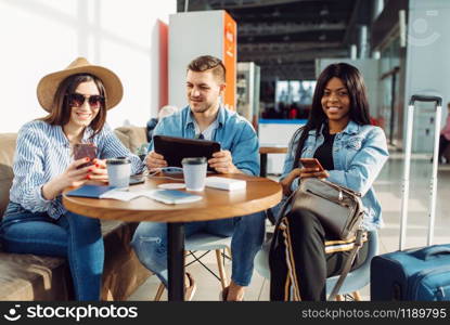 Three tourists with phones and laptop at the table waiting for departure in airport. Passengers with baggage look forward to flight in air terminal, happy journey, summer travel on vacation