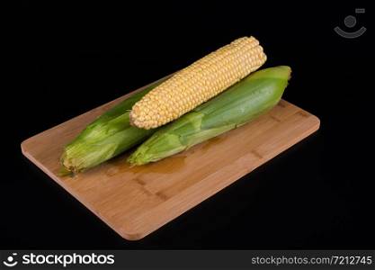 three unpeeled corn lie on a wooden board for cooking, background is black, top view. three corn in husk