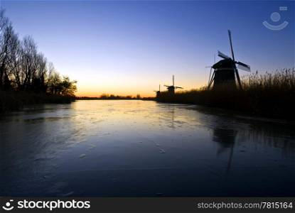 Three windmills at an early morning winter sunrise, reflecting in the cracked ice of a canal