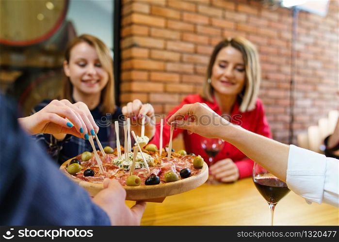 Three women caucasian girls sitting by the table holding glasses of red wine while unknown man is serving appetizer friends smiling in day at home or restaurant
