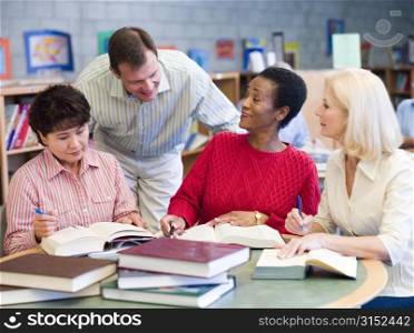Three women sitting in library with books and notepads while a man leans over them (selective focus)