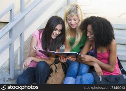 Three women sitting on staircase outdoors looking at notebook (high key)