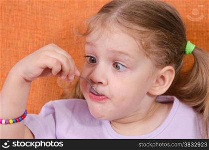 Three year old girl with pleasure and humor eats honey spoon sitting on the couch