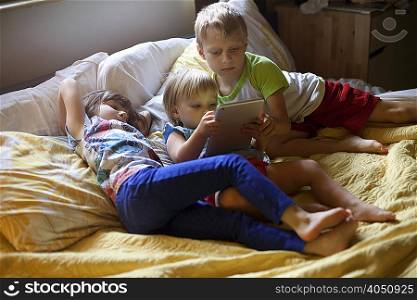 Three young children relaxing on bed using digital tablet