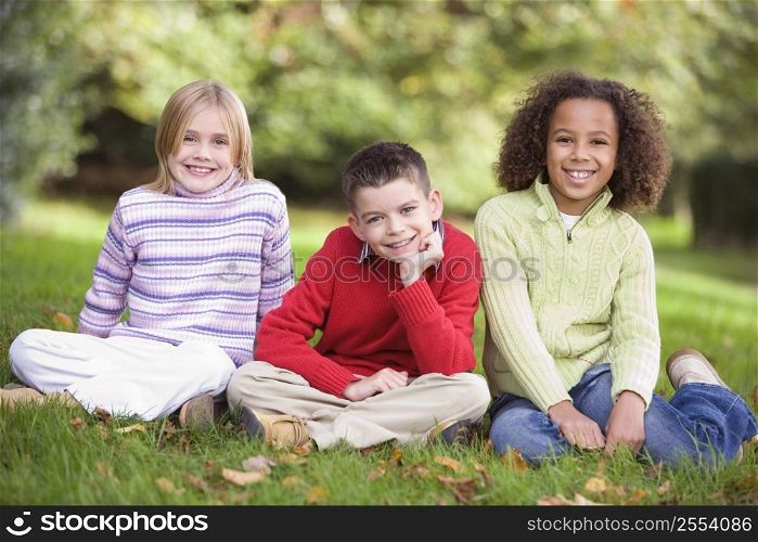 Three young children sitting outdoors in park smiling (selective focus)