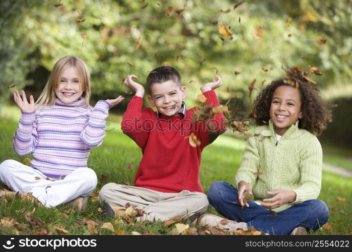 Three young children sitting outdoors in park throwing leaves in air and smiling (selective focus)