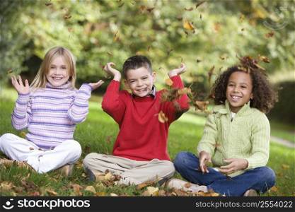 Three young children sitting outdoors in park throwing leaves in air and smiling (selective focus)