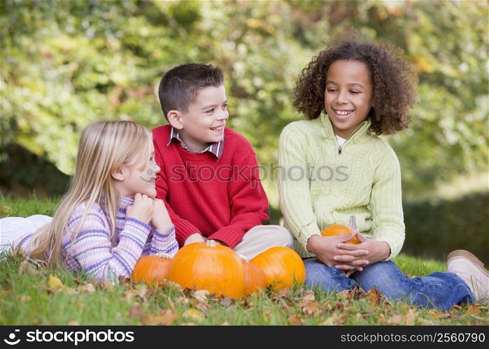 Three young friends sitting on grass with pumpkins smiling
