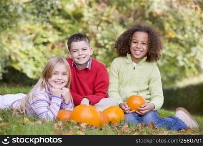 Three young friends sitting on grass with pumpkins smiling