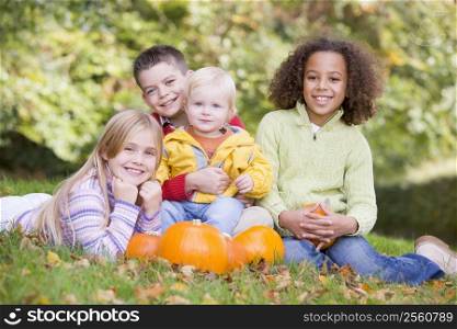 Three young friends with baby sitting on grass with pumpkins smiling
