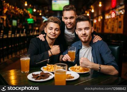 Three young men poses at the table with beer, crisps and crackers, sport bar interior on background, happy friendship of football fans
