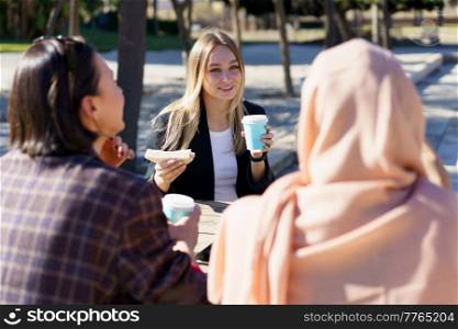 Three young multiethnic ladies friends in casual clothes smiling while resting in outdoor cafe and drinking coffee from disposable cups. Three diverse girls having coffee break and chatting in city park