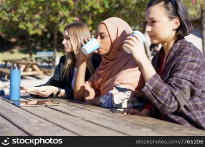 Three young multiethnic ladies friends in casual clothes smiling while resting in outdoor cafe and drinking coffee from disposable cups. Three diverse girls having coffee break and chatting in city park