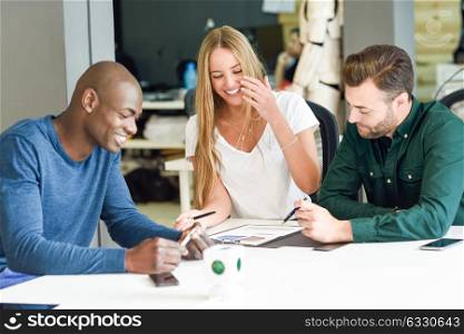 Three young people studying with graphics on white table. Beautiful blonde girl, african and caucasian men working together wearing casual clothes and smiling. Multi-ethnic group.