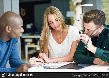 Three young people studying with graphics on white table. Beautiful blonde girl, african and caucasian men working together wearing casual clothes. Multi-ethnic group.