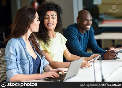 Three young people studying with laptop computer on white desk. Beautiful girls and man working toghether wearing casual clothes. Multi-ethnic group.