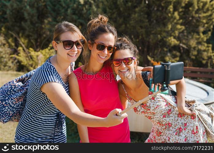 Three young woman tourists using selfie stick and mobile smart phone to make self portrait in sunny autumn day wearing summer dress
