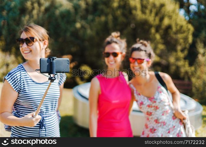 Three young woman tourists using selfie stick and mobile smart phone to make self portrait in sunny autumn day wearing summer dress