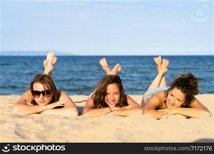 Three young women girl friends or sisters laying on the sand beach by the sea or ocean having fun in summer day sunny sun tanning wearing bikini swimsuit happy tourist on the vacation
