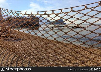 Through pattern of large cells of the fishing net, drying in the sun, a pier, an old castle and the coastline of the sea can be seen.. Fishing net dries in the sun and is blown by the wind, stretched against the backdrop of the sea coast.