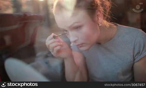 Through the window portrait of a beautiful girl applying mascara to her eyelashes.