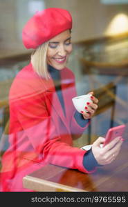 Through window of positive female in red coat and beret text messaging on cellphone while sitting with cup of coffee in cafeteria. Through glass of woman browsing smartphone in cafe