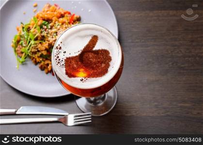 Thumbs up silhouette on foam in beer glass on black table with food, view from above.