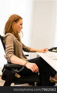 Tied to the office. Tied to the office - Young beautiful business woman strapped to chair with handcuffs in front of her computer in a modern office