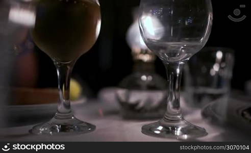 Tilt close-up shot of pouring white wine into the glass, full one standing nearby. Serving in restaurant